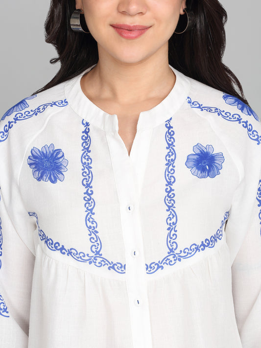 Close-up of round neck, blue floral embroidery, smocked cuffs, and gather detailing on white linen blend shirt